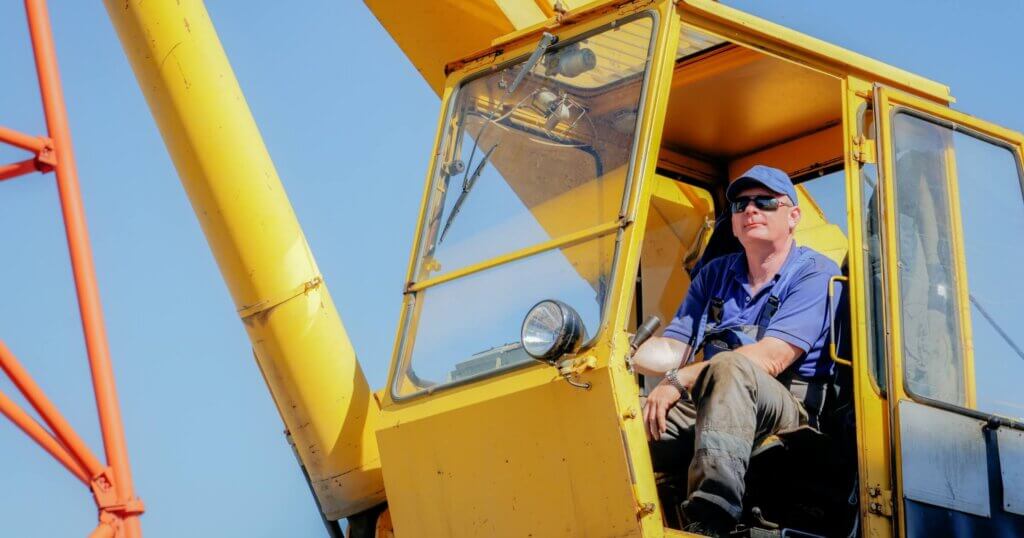 A construction worker operating a yellow crane on site, wearing a blue shirt and hard hat, illustrating machinery control, operations, and construction work.