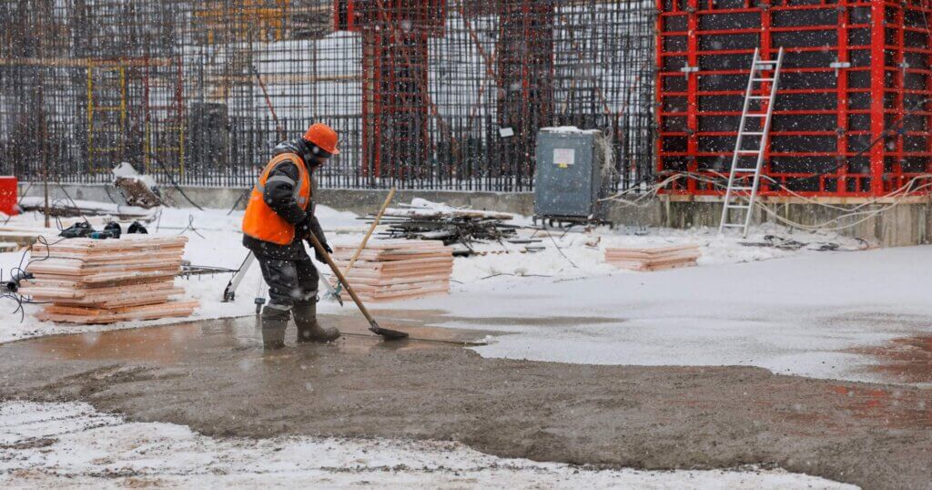 A worker in overalls is leveling concrete at a construction site with a shovel. Working in winter in a snowstorm and snow.