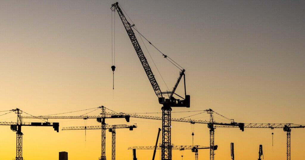 A series of construction cranes rise against a colorful sunset, casting long shadows over a busy construction site, signaling activity in the early evening light.