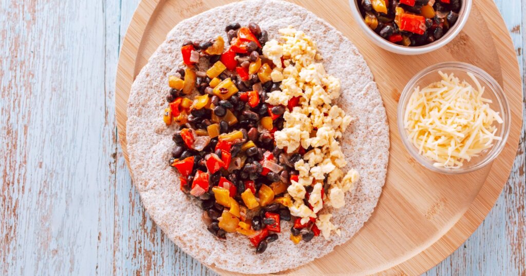 Healthy high protein burritos with eggs, bell peppers, sweet potato, black beans on whole wheat tortillas with grated cheese, top view. Healthy breakfast on wooden table.