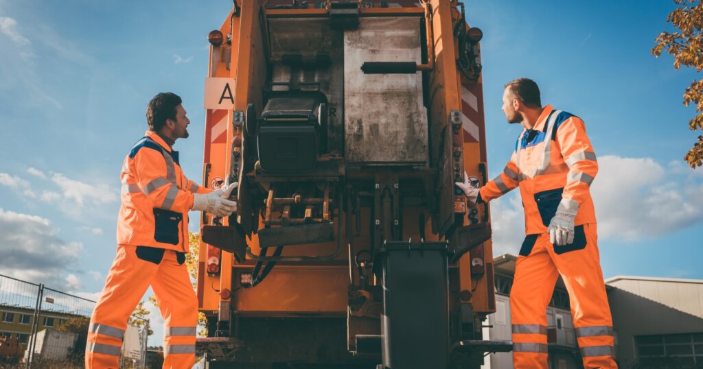 Two refuse collection workers loading garbage into waste truck emptying containers.