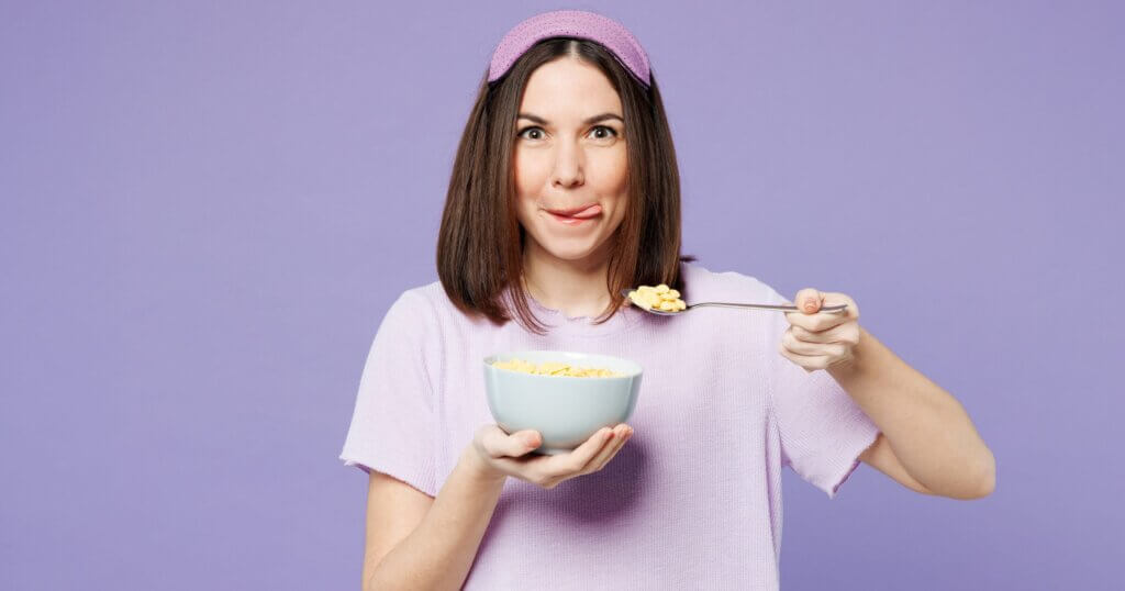 Young happy woman wear pyjamas jam sleep eye mask rest relax at home eat breakfast muesli cereals with milk fruit in bowl isolated on plain pastel light purple background. Good mood night nap concept.