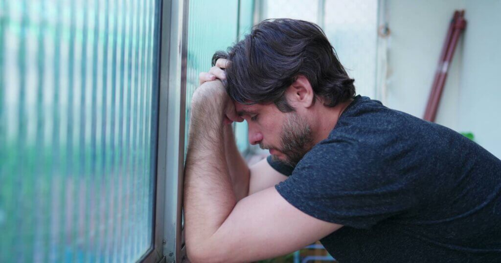Stressed man struggling with mental illness leaning by window suffering from emotional despair. One worried male person in 30s feeling pressure.