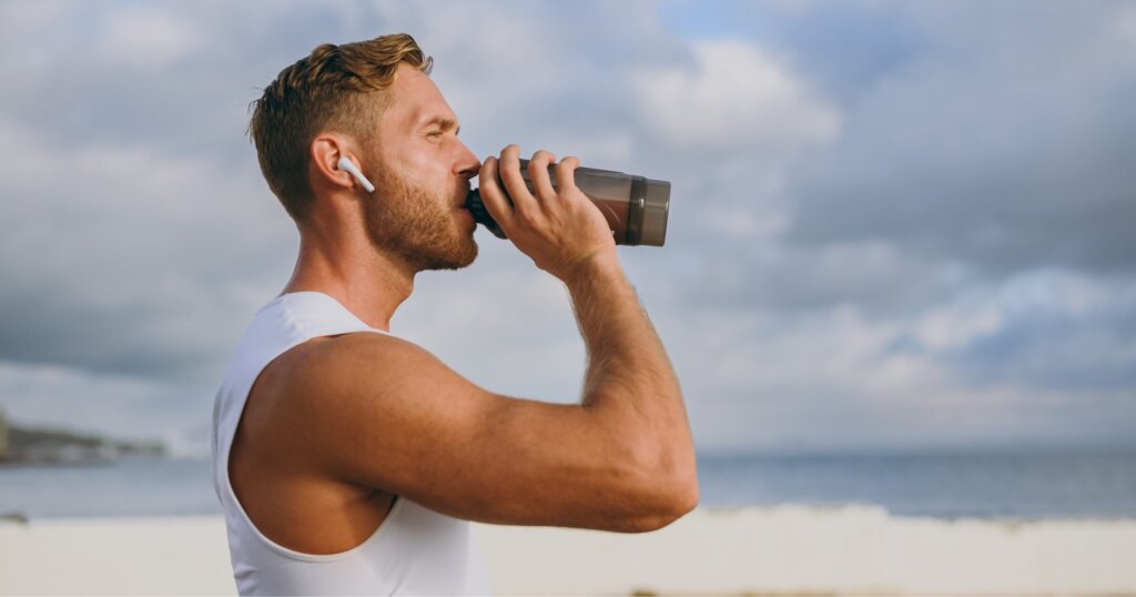 Sideways young strong athletic toned fit sportsman man he wear sports clothes earphones drink protein shake warm up train at sunrise sun dawn over sea beach outdoor on seaside in summer day morning.