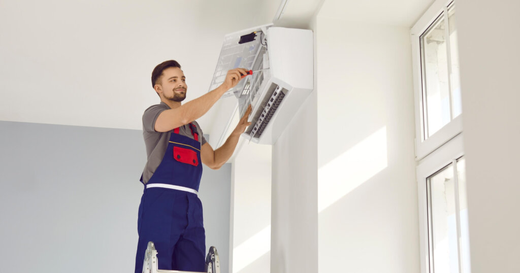 Male worker fixing air conditioning at home. Man in uniform standing on ladder stairs and installing new air conditioner on white wall in house. AC installation, maintenance, repair service concept