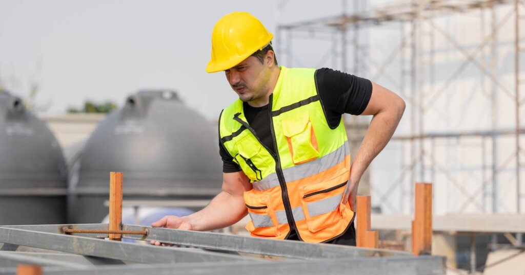 Construction worker in safety vest and hard hat inspecting structure at building site, experiencing low back pain. Concept of occupational health and safety.