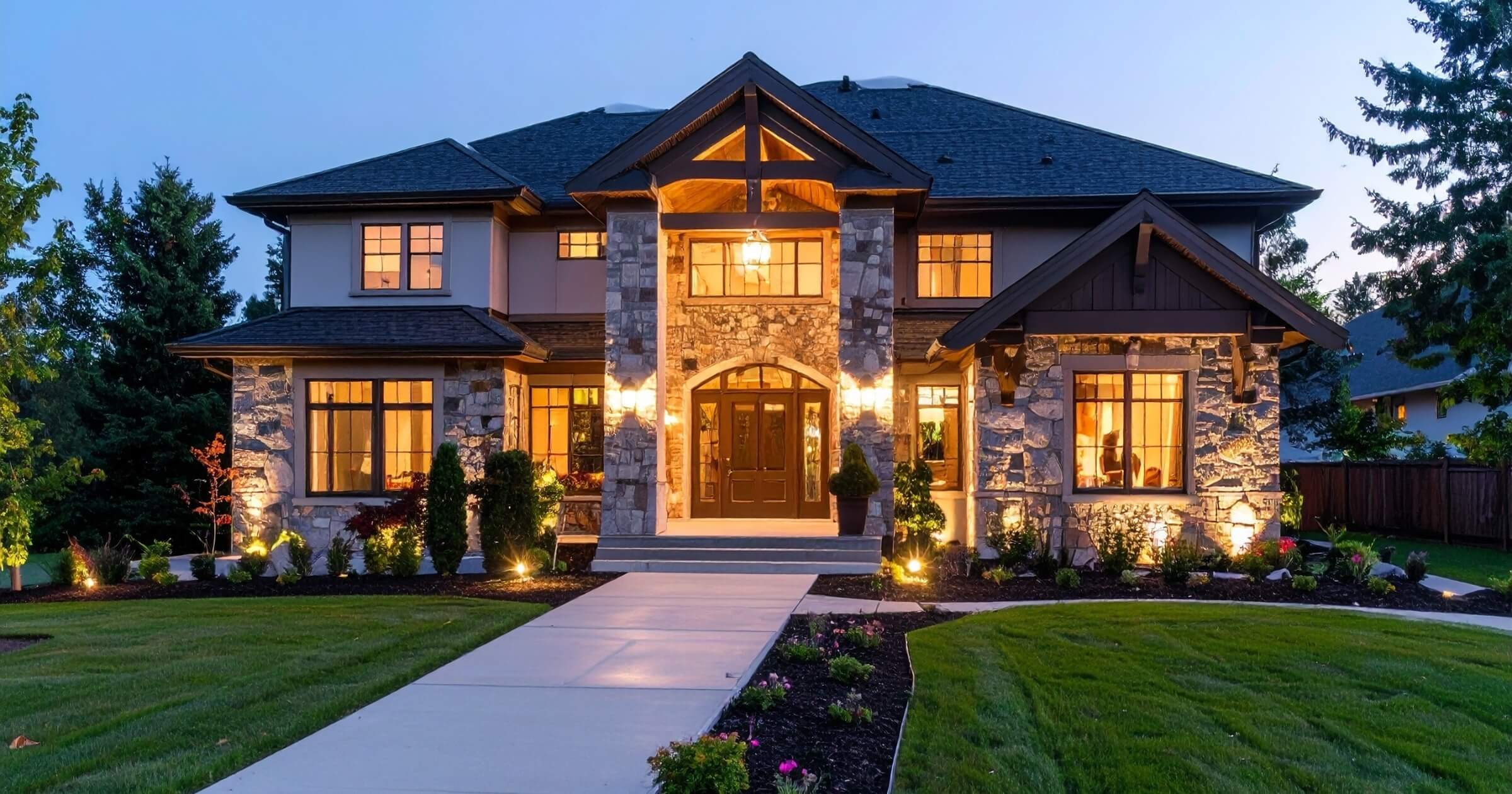 A luxurious twostory house, illuminated at dusk, features stone facade, large windows, landscaped lawn, and a paved walkway leading to the grand entrance.
