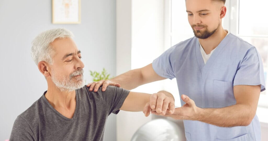 Young male physiotherapist helps old white haired retired man regain elbow mobility.