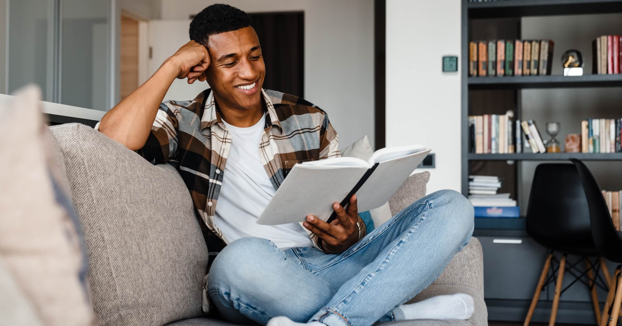 Young positive smiling african man wearing plaid shirt reading book while sitting on couch at home