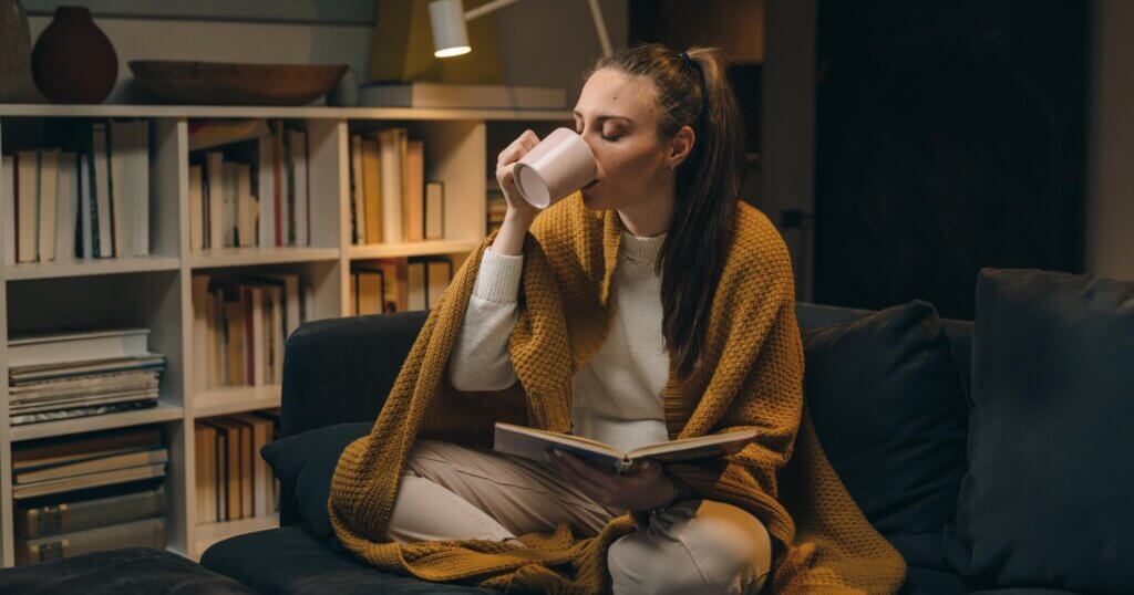 young woman reading book while sitting on sofa at her home. she is covered with blanket and drinking tea