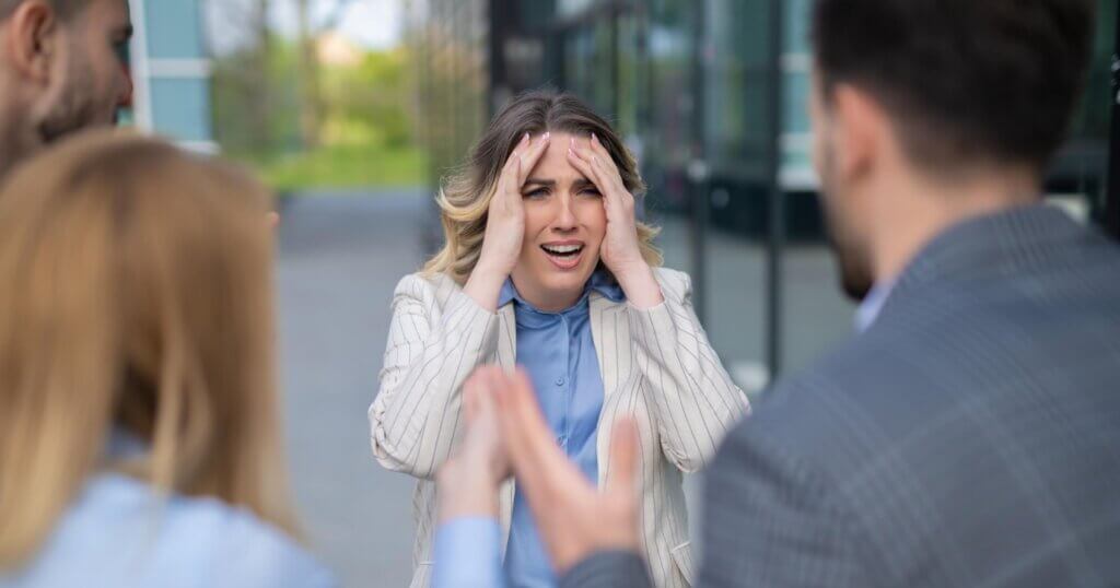 Distressed woman in striped blazer covering her head with hands, surrounded by coworkers in argument, highlighting toxic work culture and mental health issues.