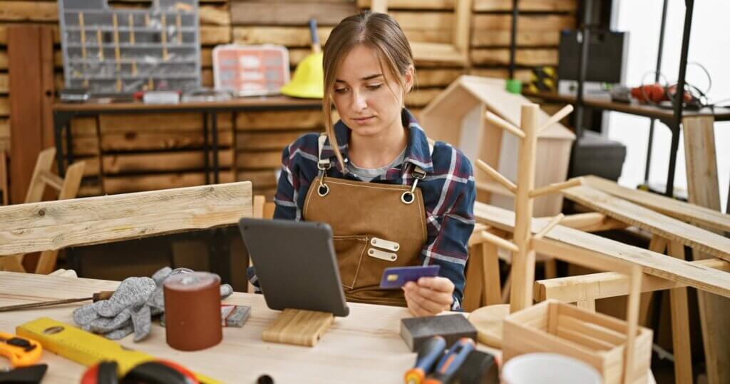 Attractive young blonde female carpenter, engrossed in serious business of financing her carpentry workshop, using credit card touchpad amidst timber, indoor.