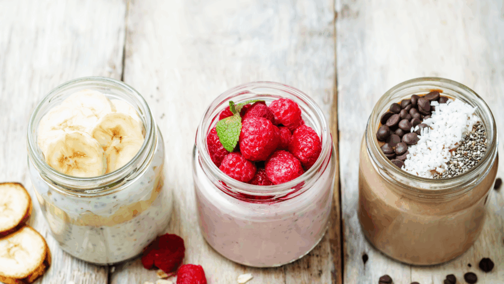 Three mason jars of overnight oats recipe variations with banana slices, fresh raspberries, and chocolate chips with coconut and chia seeds on a wooden surface